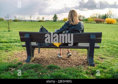 Glückliche blonde Frau in Stil Kleidung ruhen, entspannen auf der Bank sitzen und genießen Sie in der Stadt grünen Naturpark im Freien. Rückansicht Stockfoto