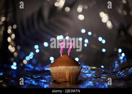 Digitales Grußkartenkonzept für Urlaub oder Geburtstag. Leckerer Vanillekuchen oder Muffin mit der Nummer 77 siebenundsiebzig auf Aluminiumfolie und verschwommenem dunklen Hintergrund. Hochwertige Fotos Stockfoto