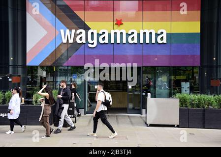 Die Menschen gehen am Wagamama Restaurant am St. Peter's Square, Manchester, England, Großbritannien, den Britischen Inseln vorbei, Jetzt mit der Fortschrittsflagge auf der Vorderseite. Die Progress Pride Flagge wurde 2018 vom Künstler Daniel Quasar entwickelt. Das Redesign basiert auf der legendären Regenbogenflagge von 1978 und feiert die Vielfalt der LGBTQ-Gemeinschaft und fordert eine integrativere Gesellschaft. Stockfoto