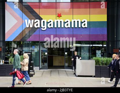 Die Menschen gehen am Wagamama Restaurant am St. Peter's Square, Manchester, England, Großbritannien, den Britischen Inseln vorbei, Jetzt mit der Fortschrittsflagge auf der Vorderseite. Die Progress Pride Flagge wurde 2018 vom Künstler Daniel Quasar entwickelt. Das Redesign basiert auf der legendären Regenbogenflagge von 1978 und feiert die Vielfalt der LGBTQ-Gemeinschaft und fordert eine integrativere Gesellschaft. Stockfoto