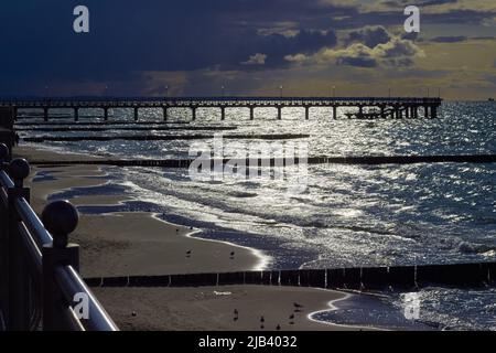 Promenade und Wellenbrecher in der untergehenden Sonne. Möwen am Strand. Helle Lichter und tiefe Schatten. Stockfoto