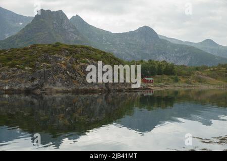 Norwegische Meereslandschaft, wilde Hütte am Meer, felsige Küste mit dramatischem Himmel, die Sonne bricht durch die Wolken, schiere Klippen, kleine Inseln beleuchtet Stockfoto