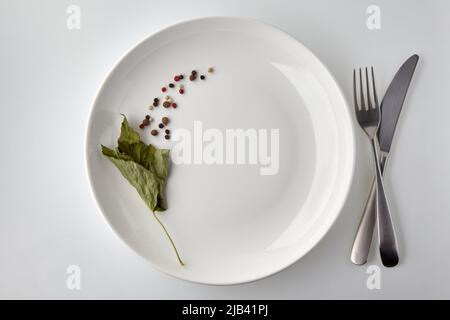 Weißer Teller mit verschiedenen Pfefferkörnern und trockenen Johannisbeerblättern auf weißem Hintergrund. Speisekarte im Restaurant. Draufsicht Stockfoto