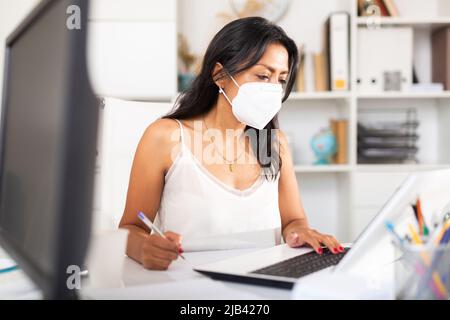 Latina in einer medizinischen Maske, die im Büro am Laptop arbeitet Stockfoto