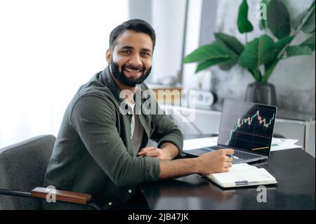 Porträt eines attraktiven positiven indischen oder arabischen Krypto-Investors, Börsenmaklers, Kryptowährungsexperten, sitzen an einem Schreibtisch im modernen Büro, blicken freundlich lächelnd auf die Kamera Stockfoto