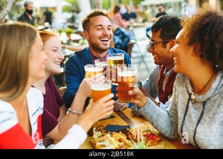 Eine Gruppe von begeisterten multirassischen Freunden, die sich gegenseitig angucken, während sie mit Biergläsern am Tisch mit Essen auf der Terrasse des Pubs jubeln Stockfoto