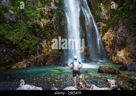 Ein Mann in einem Hut bleibt unter den Strömen des Wasserfalls. Der Grunas Wasserfall ist ein malerischer Ort im Nationalpark Théthis, Albanien Stockfoto