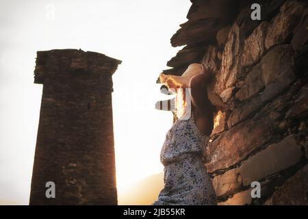 Ein junges Mädchen in Kleid und Hut steht bei Sonnenuntergang neben den mittelalterlichen Türmen in Svaneti. Stockfoto