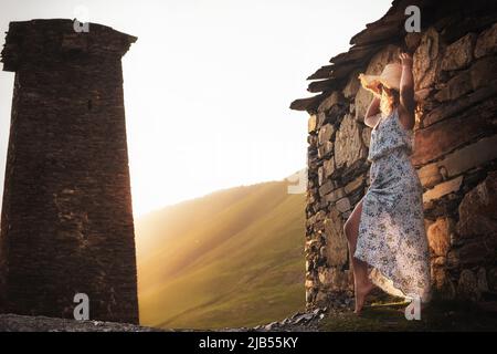 Ein junges Mädchen in Kleid und Hut steht bei Sonnenuntergang neben den mittelalterlichen Türmen in Svaneti. Stockfoto