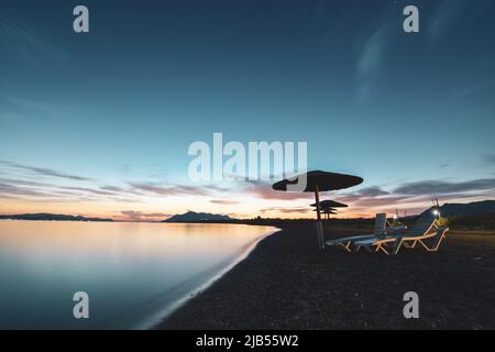 Leere Sonnenliegen und Sonnenschirme am Sandstrand bei Mondschein in der Nacht in Agean Sea, Griechenland Stockfoto