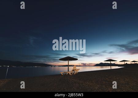 Leere Sonnenliegen und Sonnenschirme am Sandstrand bei Mondschein in der Nacht in Agean Sea, Griechenland Stockfoto
