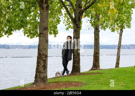 Lächelndes biracial Teenager-Mädchen oder junge Erwachsene Frau in grauer Jacke lehnt sich an Baum im Park am See Stockfoto