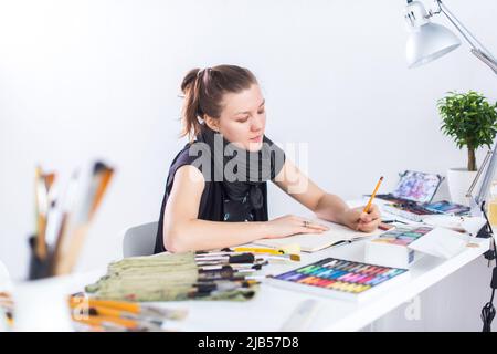 Junge Künstlerin zeichnet Skizze mit Skizzenbuch mit Bleistift an ihrem Arbeitsplatz im Studio. Seitenansicht Porträt eines inspirierten Malers Stockfoto