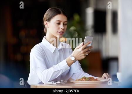 Content junge Geschäftsfrau in weißer Bluse sitzt mit Gericht am Tisch und überprüft Messenger auf dem Smartphone, während sie Business Lunch im Restaurant Stockfoto