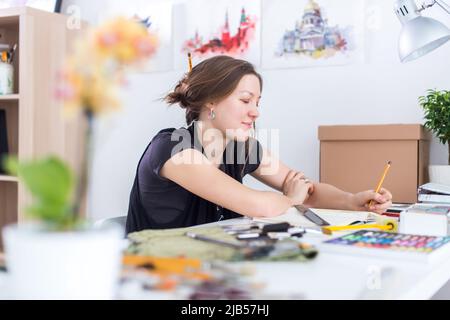 Junge Künstlerin zeichnet Skizze mit Skizzenbuch mit Bleistift an ihrem Arbeitsplatz im Studio. Seitenansicht Porträt eines inspirierten Malers Stockfoto