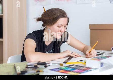 Junge Künstlerin zeichnet Skizze mit Skizzenbuch mit Bleistift an ihrem Arbeitsplatz im Studio. Seitenansicht Porträt eines inspirierten Malers Stockfoto