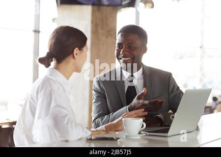 Fröhliche, aufgeregte junge, multiethnische Geschäftspartner in formalwear, die an der Theke sitzen und einen Laptop benutzen, während sie über Zusammenarbeit diskutieren Stockfoto