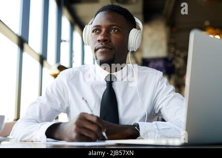 Ernst nachdenklicher junger afroamerikanischer Manager in weißem Hemd und Krawatte, der Musik in Kopfhörern hört und wegschaut, während er eine Idee für BU findet Stockfoto