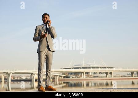Lächelnder, selbstbewusster junger afroamerikanischer Geschäftsmann in modischem Anzug, der sich gegen das Stadtbild stellt und Arbeitsprobleme am Telefon löst Stockfoto