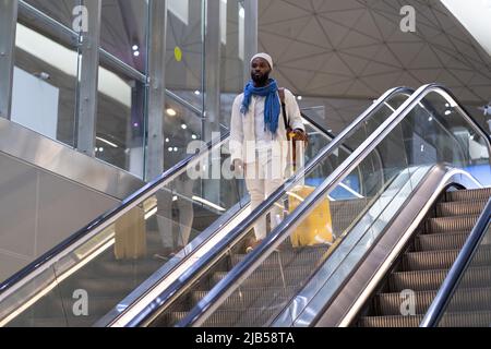 Afroamerikanischer Passagiermann mit Koffer steht auf Rolltreppe, hält Handlauf im Flughafenterminal Stockfoto