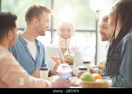 Gruppe von fröhlichen jungen multiethnischen Freunden in legerer Kleidung, die an einem Tisch sitzen und Pizza essen, während sie sich während des Mittagessens unterhalten Stockfoto