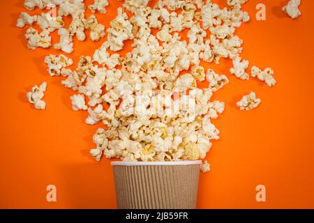 Popcorn wird aus dem Glas auf einem orangefarbenen Hintergrund ausgestreut, von oben betrachtet. Ein Spritzer Popcorn Stockfoto