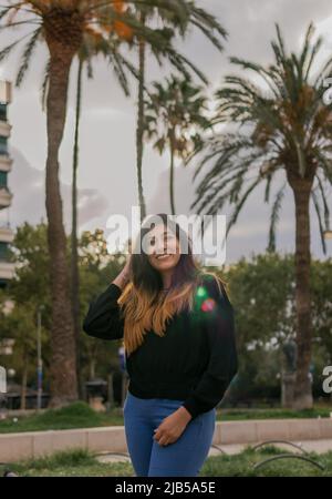 Eine Gegenlicht-Fotografie mit einem Lichtreflexe einer lächelnden hispanischen Frau, die auf einer Straße steht, wobei ihre rechte Hand ihren Kopf und Palmen im b berührt Stockfoto