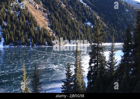 Kolsai See im Eis mit Bäumen und Berghang im Winter bei sonnigem Wetter Stockfoto