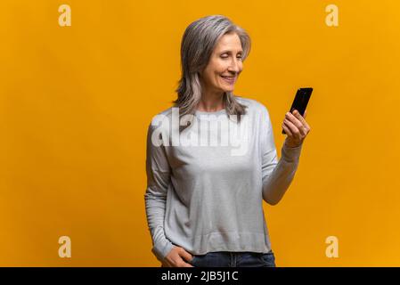 Profilportrait der glücklichen trendigen reifen Dame mit Smartphone und lächelnd, zufrieden mit der Verbindungsqualität. Innenaufnahme des Studios isoliert auf gelbem Hintergrund Stockfoto
