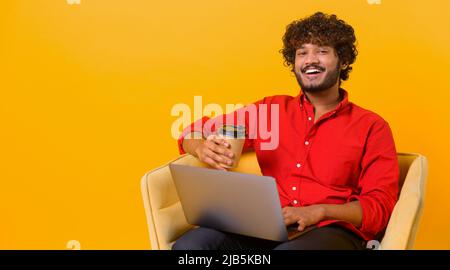 Ein geschäftiger und selbstbewusster Mann aus dem Nahen Osten trägt ein elegantes, legeres Hemd, das auf dem Laptop SMS schreibt und eine Tasse Kaffee zum Mitnehmen hält. Männlicher Büroangestellter auf Orange isoliert Stockfoto