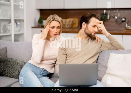 Traurige Frau, die auf den Laptop-Bildschirm schaut und sich Sorgen um die Ausgaben zu Hause macht. Ehepartner sitzen zu Hause auf der Couch mit einem Laptop. Unglaubliche Nachrichten oder der Gewinn der Lotterie hat das Paar begeistert Stockfoto