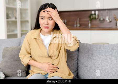 Junge asiatische Frau, die auf dem Sofa sitzt und an Magenschmerzen leidet, die Hände zu Hause auf dem Bauch hält, auf Arzt, Vergiftung, Menstruation wartet Stockfoto