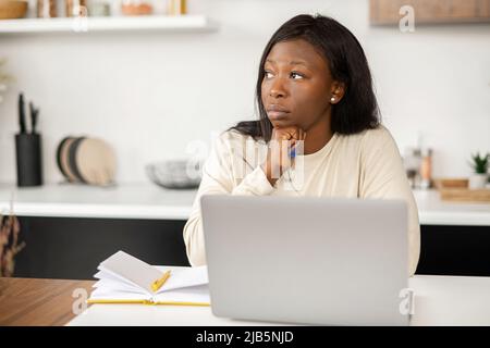 Besorgte afroamerikanische Frau sitzt mit einem Laptop in der Küche zu Hause, traurig wegen empfangener Ablehnung, müde von der Online-Arbeit, junge Frau fühlt sich deprimiert Stockfoto