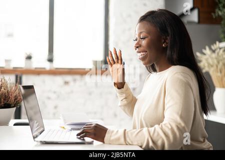 Afroamerikanische Frau sitzt drinnen und nutzt die App auf dem Laptop für Videoanruf an Kollegen, Kollegen oder Freunde. Junge Angestellte in Freizeitkleidung, die online über Videoverbindung spricht Stockfoto