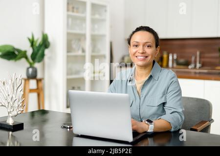 Weibliche Angestellte in eleganter Freizeitkleidung blickt auf die Kamera und lächelt, tippt Nachrichten, beantwortet E-Mails, Vorderansicht. Eine selbstbewusste Frau mittleren Alters arbeitet im modernen Büro mit einem Laptop Stockfoto