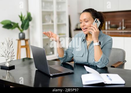 Seitenabwärts-Aufnahme einer Frau, die am Telefon spricht, während sie am Laptop arbeitet. Sie sitzt am Holztisch und lächelt, während sie mit ihrer Hand gestikuliert Stockfoto