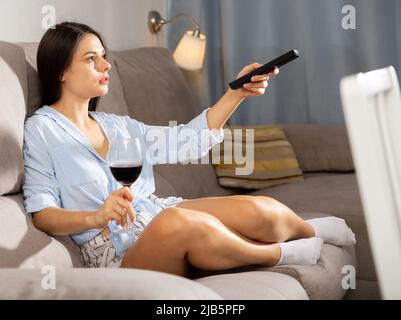 Brunette mit Fernbedienung und ein Glas Rotwein sitzt zu Hause auf dem Sofa Stockfoto