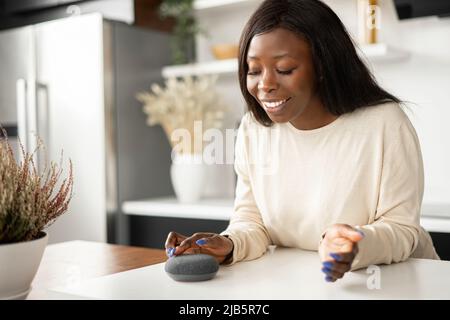 Charmante junge afroamerikanische junge Frau verwendet Sprachbefehle für drahtlose intelligente Lautsprechersteuerung. Smart Home und Technologiekonzept Stockfoto