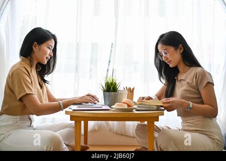 Zwei attraktive asiatische weibliche College-Studenten lesen ein Buch und machen Hausaufgaben im komfortablen Café zusammen. Rückansicht Stockfoto