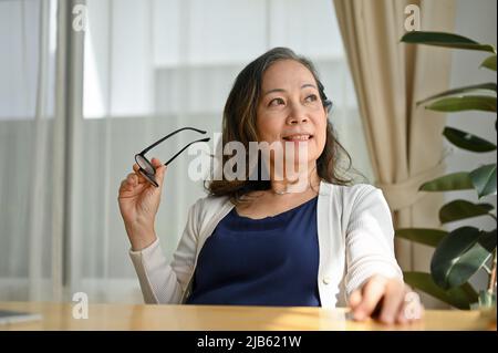 Erfolgreiche asiatische Frau, die im modernen Wohnzimmer sitzt, ihre Brille hält und mit inspiriertem und leidenschaftlichem Gefühl durch das Fenster blickt Stockfoto