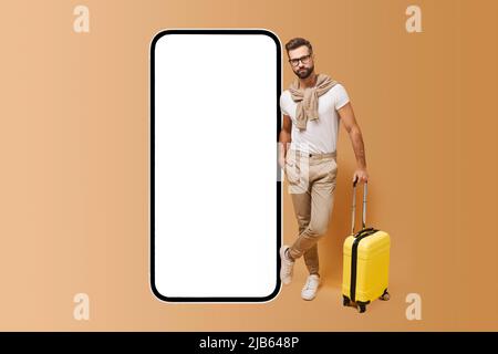 Reise-App. Schöner charismatischer bärtiger Mann steht mit Gepäck in der Nähe eines riesigen Mobiltelefons mit leerem Bildschirm, moderner Hipster-Typ in einer Brille mit gelbem Koffer, der eine App für Buchungen und Reisen anwirbt Stockfoto