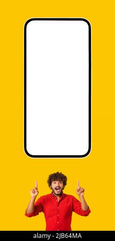 Aufgeregt erstaunt lockigen indischen bärtigen Mann zeigt die Finger nach oben auf riesige Smartphone mit leeren leeren Bildschirm isoliert auf gelb, glücklich lateinischen Kerl präsentiert mobile App, zeigt beste Deal, Kopierraum Stockfoto