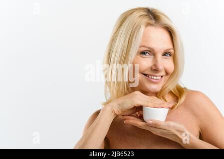 Frau mittleren Alters 50s hält Glas mit Anti-Aging-Feuchtigkeitscreme, Vorbereitung Anwendung Creme auf das Gesicht, Blick auf die Kamera isoliert auf weiß. Hautpflege Routine Konzept Stockfoto