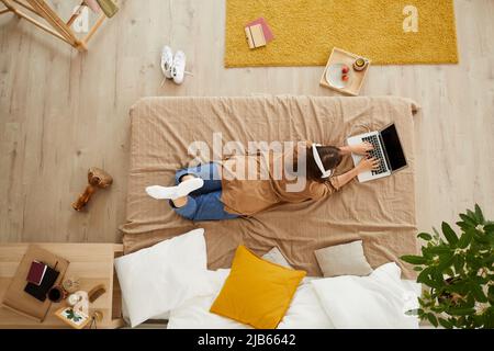 Die junge Frau in Kopfhörern, die im Bett liegt und auf dem Laptop tippt, während sie an der Forschung arbeitet, kann aus der Perspektive betrachtet werden Stockfoto