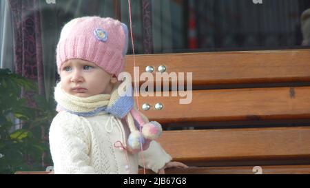 Bunt gekleidete Mädchen mit blauen Augen und bunten handgemachten Hut sitzt auf einer Bank am Straßenrand. Sie hält den fliegenden losen Faden in der Hand. Stockfoto