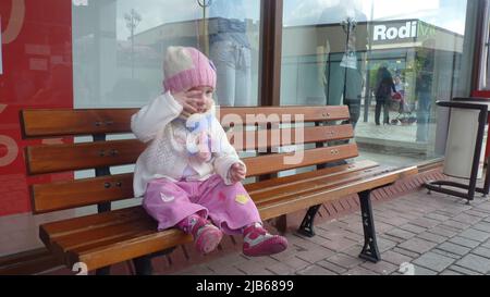 Bunt gekleidete Mädchen mit blauen Augen und bunten handgemachten Hut sitzt auf einer Bank am Straßenrand. Sie hält den fliegenden losen Faden in der Hand. Stockfoto