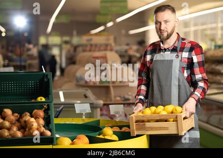 Nahaufnahme eines fröhlichen männlichen Supermarktarbeiters in guter Laune, der in einem Handkarton mit frischen Bio-Früchten hält, die Kamera anschaut und lächelt Stockfoto