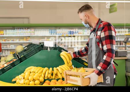 Kaukasischer Mitarbeiter im Obstladen mit Zitrusfrüchten und Bananen, Supermarktarbeiter in einer schützenden Gesichtsmaske, die Bananen aus einer Schachtel in die Regale des Ladens legt Stockfoto