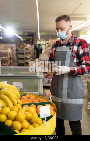 Portrait von Mitarbeitern, die digitale Tablette beim Überprüfen von Früchten in Bio-Bereich des Supermarkts, junger Mann Lebensmittelhändler in Maske Stockfoto