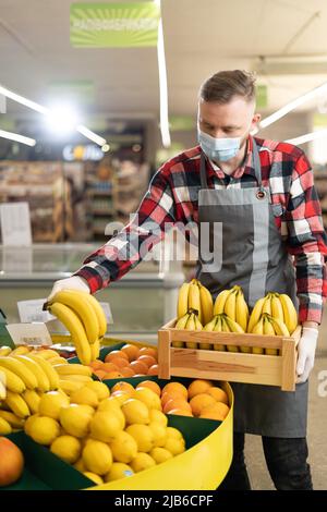 Supermarktarbeiter in grauer Schürze und Schutzmaske, die Früchte in seiner Abteilung stapeln, Mann, der Bananen in einem Geschäft verkauft Stockfoto